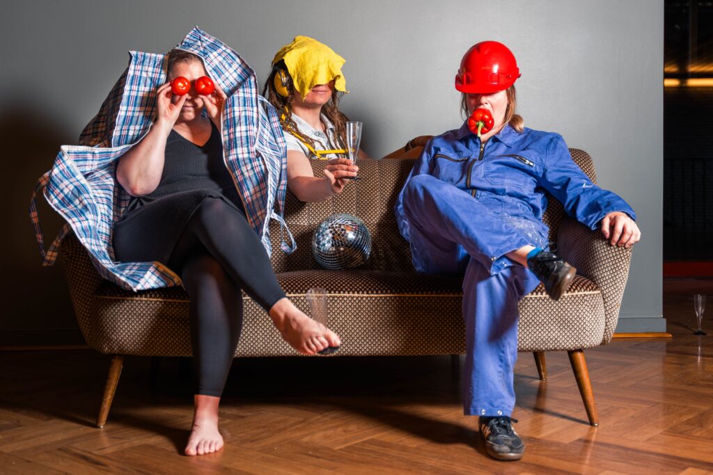 Ankündigungsfoto für Backstage Europe: Drei Personen sitzen oder lehnen auf einem Sofa, ihre Gesichter verdeckt mit unterschiedlichen Dingen: Tomaten, Wischlappen, Baustellenhelm. Dazwischen Discokugel und Sektgläser.